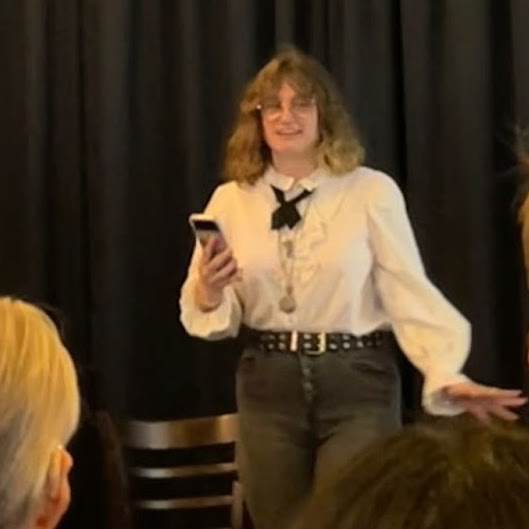 hazyn forsythe reading ‘sealed’ from monstera at a 2022 event—white ruffled shirt, black ribbon tie, layered necklaces.